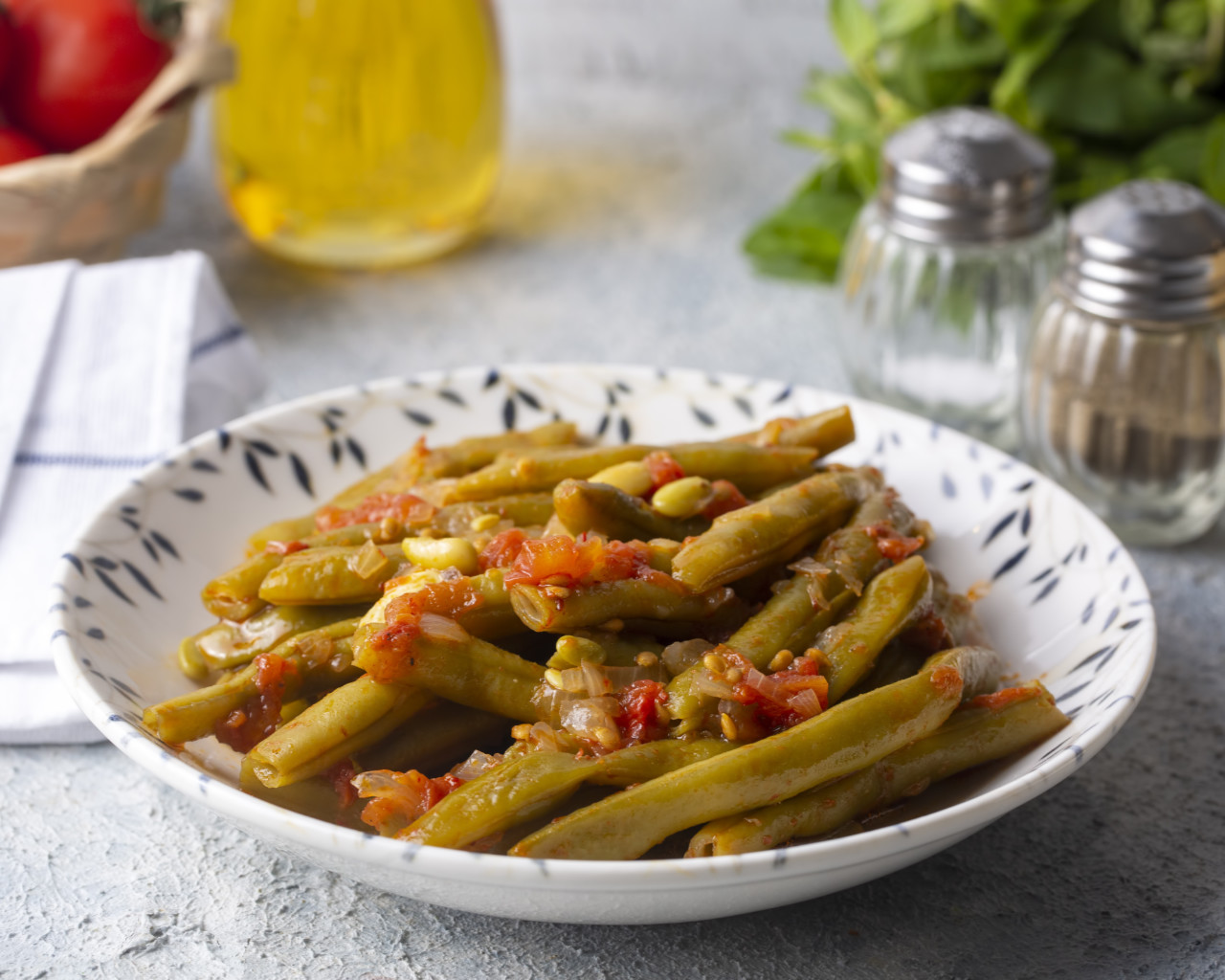 Greek fasolakia (green beans) slow-cooked in tomato sauce and olive oil, served in a patterned bowl with olive oil, tomatoes, and fresh herbs in the background