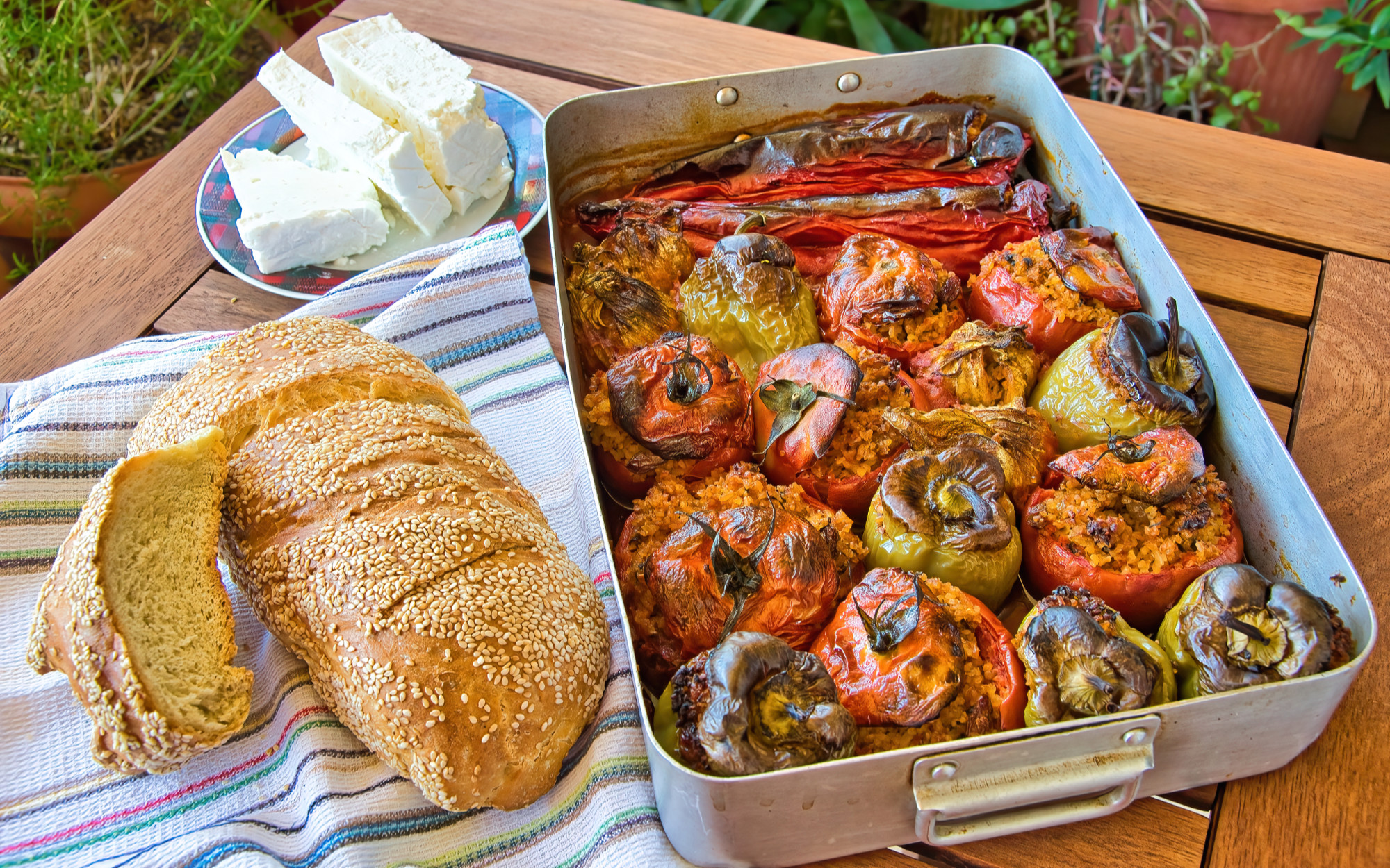 Traditional Greek gemista (stuffed tomatoes and peppers) baked in a metal pan, served with crusty sesame bread and a plate of feta cheese on a wooden table